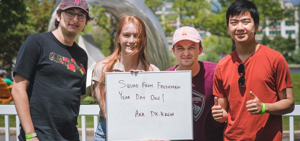 Photo of (Left to right) Andy (COM 22), Emma Nelson (COM 22), Alberto Aizenman (COM 22) and Jon Ngo (ENG 22). They stand together and hold a sign that reads "Squad from freshman year day one aka DK-Krew".