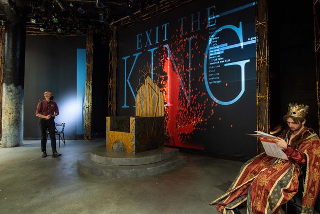 Photo the cast and crew, including artist in residence Paolo Scoppola, middle, of Exit the King running a tech rehearsal April 27 at CFA’s Studio One. A large golden throne is seen center stage; behind it, a wall that reads "EXIT THE KING" in blue and is splattered with red paint. At right, an actor, Declan Schliesmann (CFA’22), dressed as a king with red and gold robes sits and practices his lines, script in hand. Scoppola, in black pants, crimson shirt, and a face mask is seen at left.