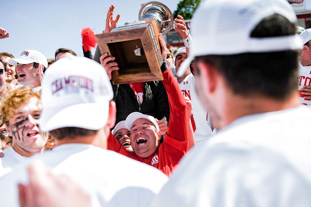 Photo of lacrosses head coach Ryan Polley hoisting the Patriot League trophy for the first time in the middle of the cheering group of lacrosse players. Polley wears a red long sleeve and players are in white. They all wear white caps.