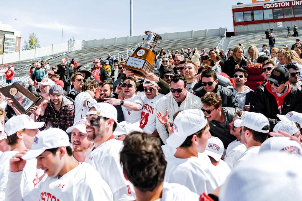 Photo of students, families, and program alumni swarming Nickerson Field and taking turns holding the Patriot League trophy. The team is seen in white and white caps as a crowd of people cheer and jump for joy around them