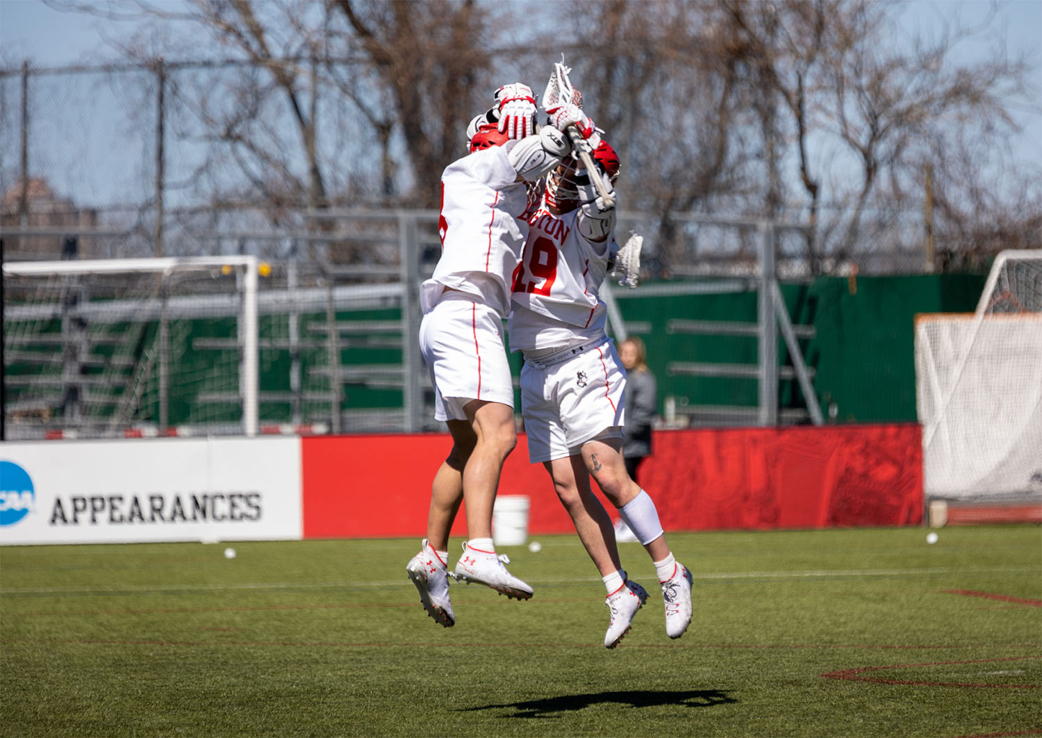 Photo of Louis Perfetto (left) and Timmy Ley jumping in the air on Nickerson field and giving each other a chest bump. Both wear white BU mens lacrosse uniforms and red helmets, with lacrosse sticks in hand. 