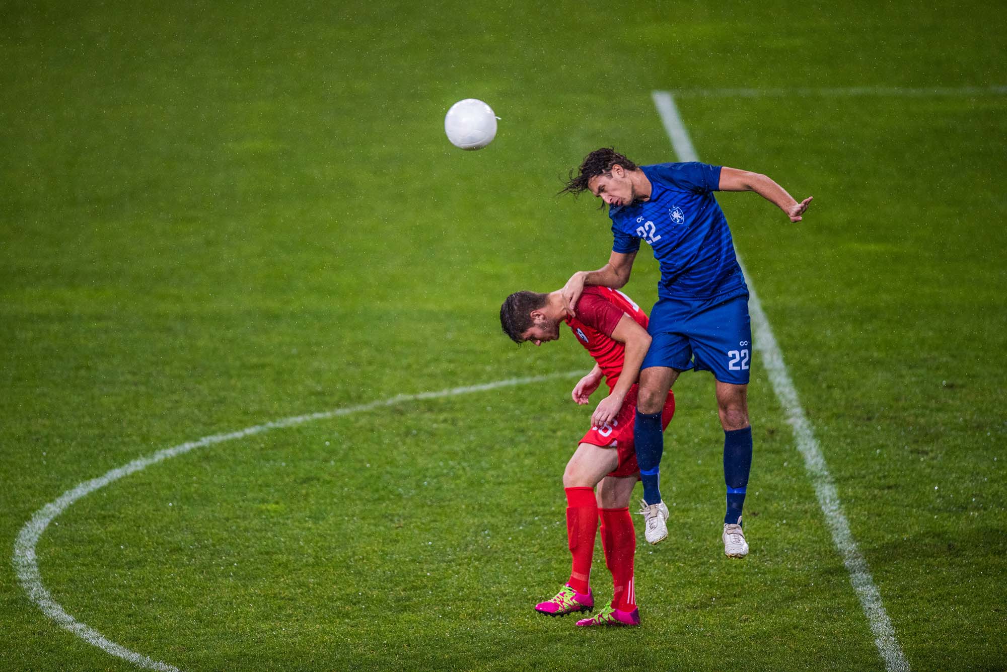 Photo of 2 soccer (football) players jumping and heading the soccer ball. A player in blue is caught mid-air after the bump, as a player in red is crouched next to him.