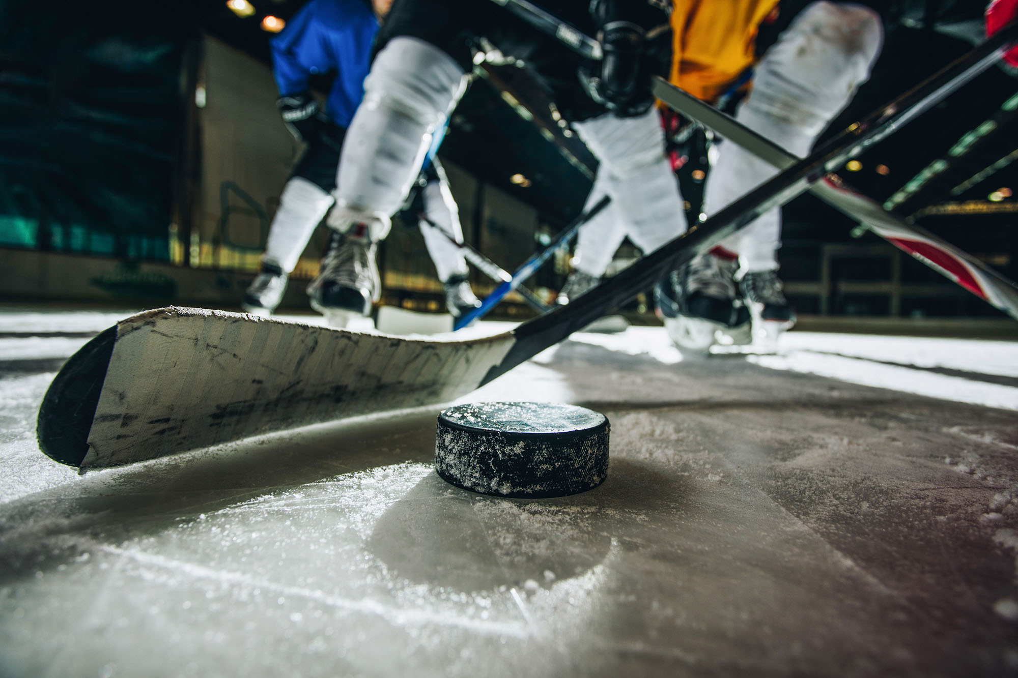 Close up photo of an ice hockey puck and stick during a match with hockey players dressed in yellow and blue uniforms in the background.