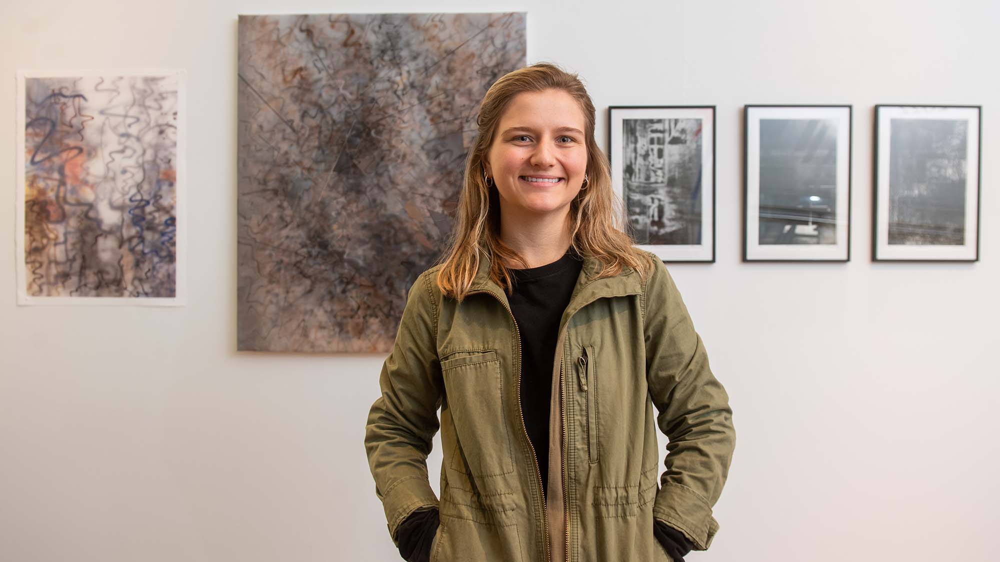 Photo of Brianna Howard (CFA’22) standing in front of her work at the BFA Thesis Art Exhibit at Stone Gallery May 5. Brianna is white woman with blonde hair; she smiles with her hands in her pockets, wears a green jacket and black plants. From left, they are named Reposition, Overlapped, Proximity, Talking with the Shadows, and Remnant. The prints are mostly gray and tan colors and are abstract in nature, featuring long curving lines.