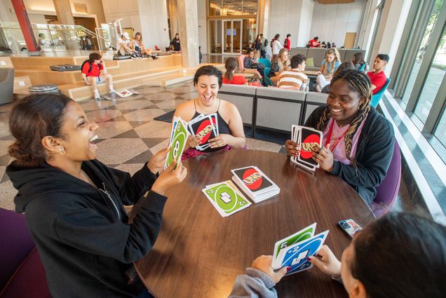 Photo of 4 incoming students playing Uno with a set of giant Uno cards. They sit around a round wooden table as they laugh and hold the giant cards in their hands. A draw pile of cards can be seen in the middle of the table.