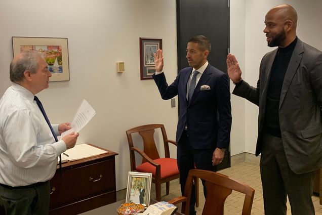 Photo of Alex Geourntas, Interim City Clerk at City of Boston, swearing in Sandro Galea (left), a Latino man wearing a navy suit and raising his right arm, and Greg Wilmot, a tall Black man wearing a navy turtleneck and black blazer and pants who is also raising his right arm, at Boston City Hall on June 3.