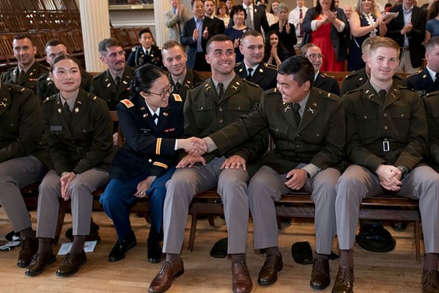 Photo of ROTC cadets sitting in pews wearing dark olive green uniform tops and grey slacks. One woman in the middle wears navy slacks. They smile and pose for the camera. Two in the front row reach over a cadet in the middle and shake hands.