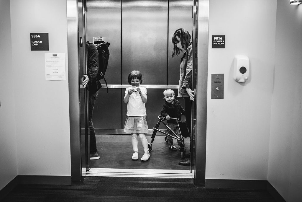 A family of four on a modern elevator. One child is wearing a white shirt and skirt and the other is wearing a shirt and pants while using a walker to assist with mobility