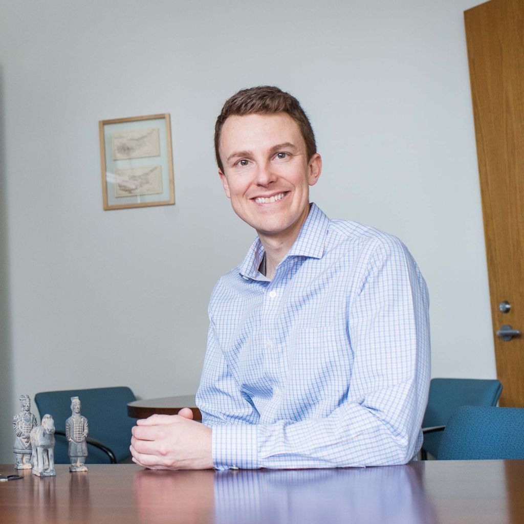 John M. Marston sits and smiles at the camera in a blue collared shirt. There is a wooden desk and bookcase nearby.