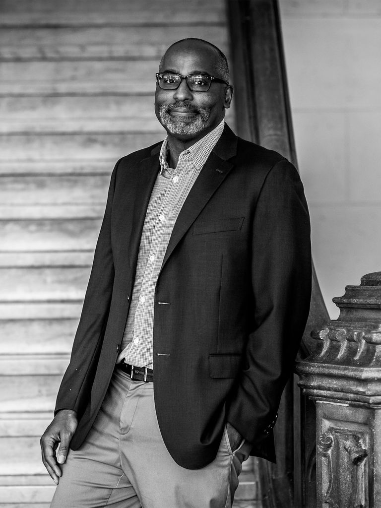 Harvey Young smiles at the camera while standing on a staircase in a suit jacket in this black and white photo