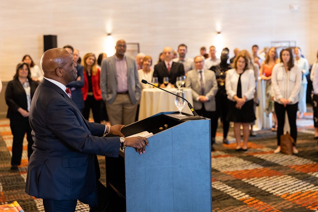 Kenneth Elmore stands before a group of his colleagues at a going away party at the Hotel Commonwealth on June 22nd, 2022. Elmore is wearing a dark suit and stands at a podium in a well lit ballroom.