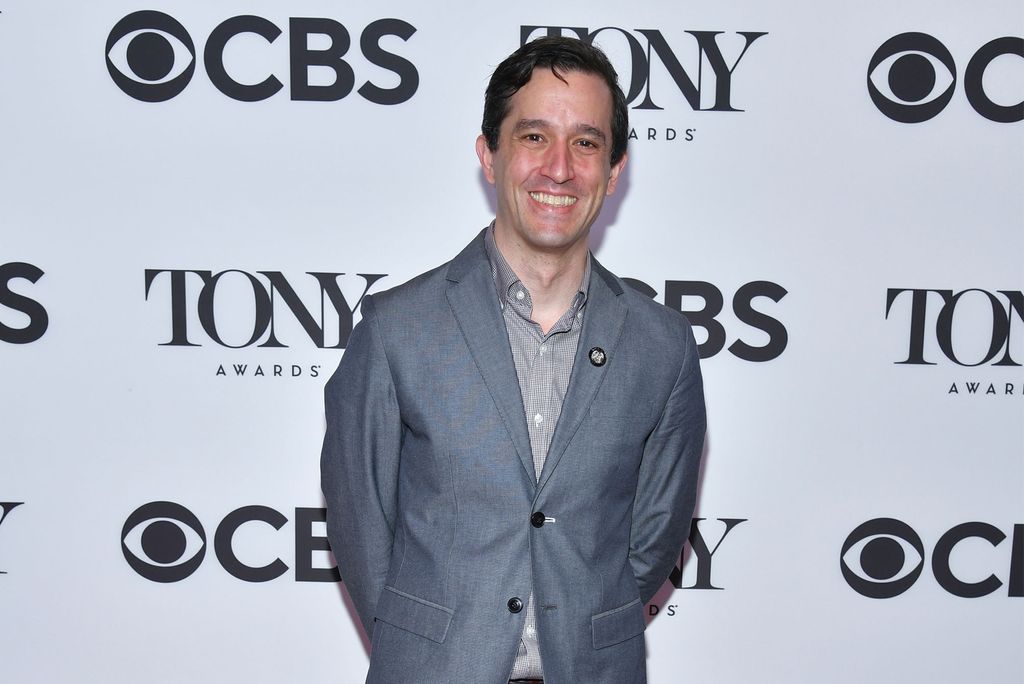 A photo of Drew Levy in a Grey Suit at an event with a CBS and Tony Award photo board behind him