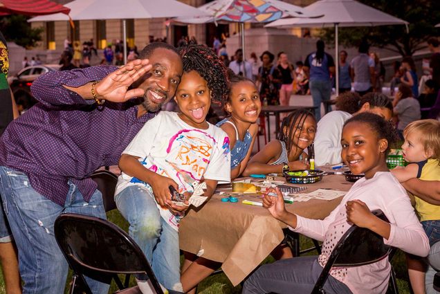 Photo of a family smiling and posing at an outdoor table during a the evening. A black man waves tot he camera as he hugs a young black girl next to him. Other black children sit around a table with a tan tablecloth and smile.