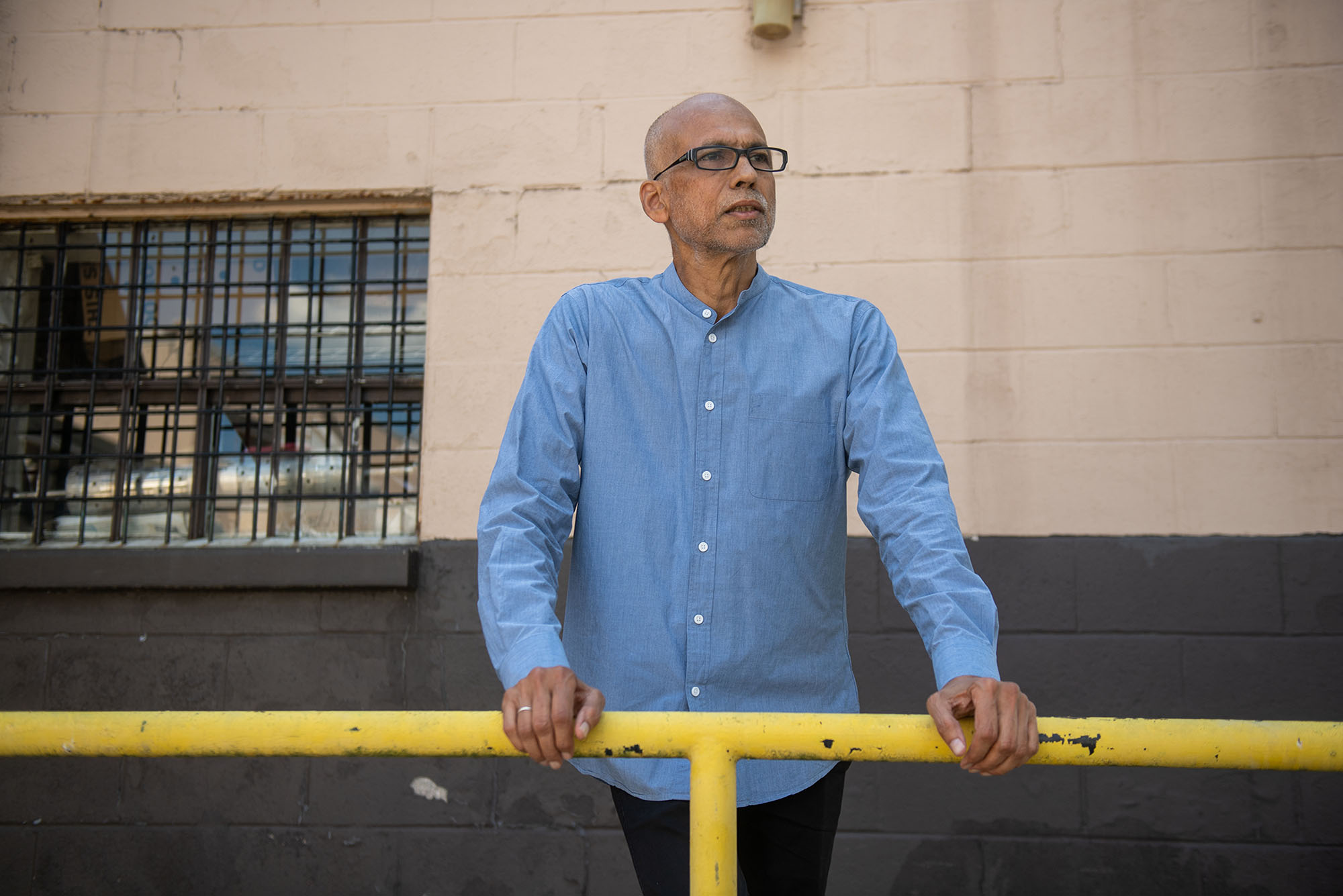 photo of André de Quadros, choir director and teacher of music in the BU prison education program, looking at the camera with a stoic expression. He wears black, rectangular glasses and a light blue collared shirt.