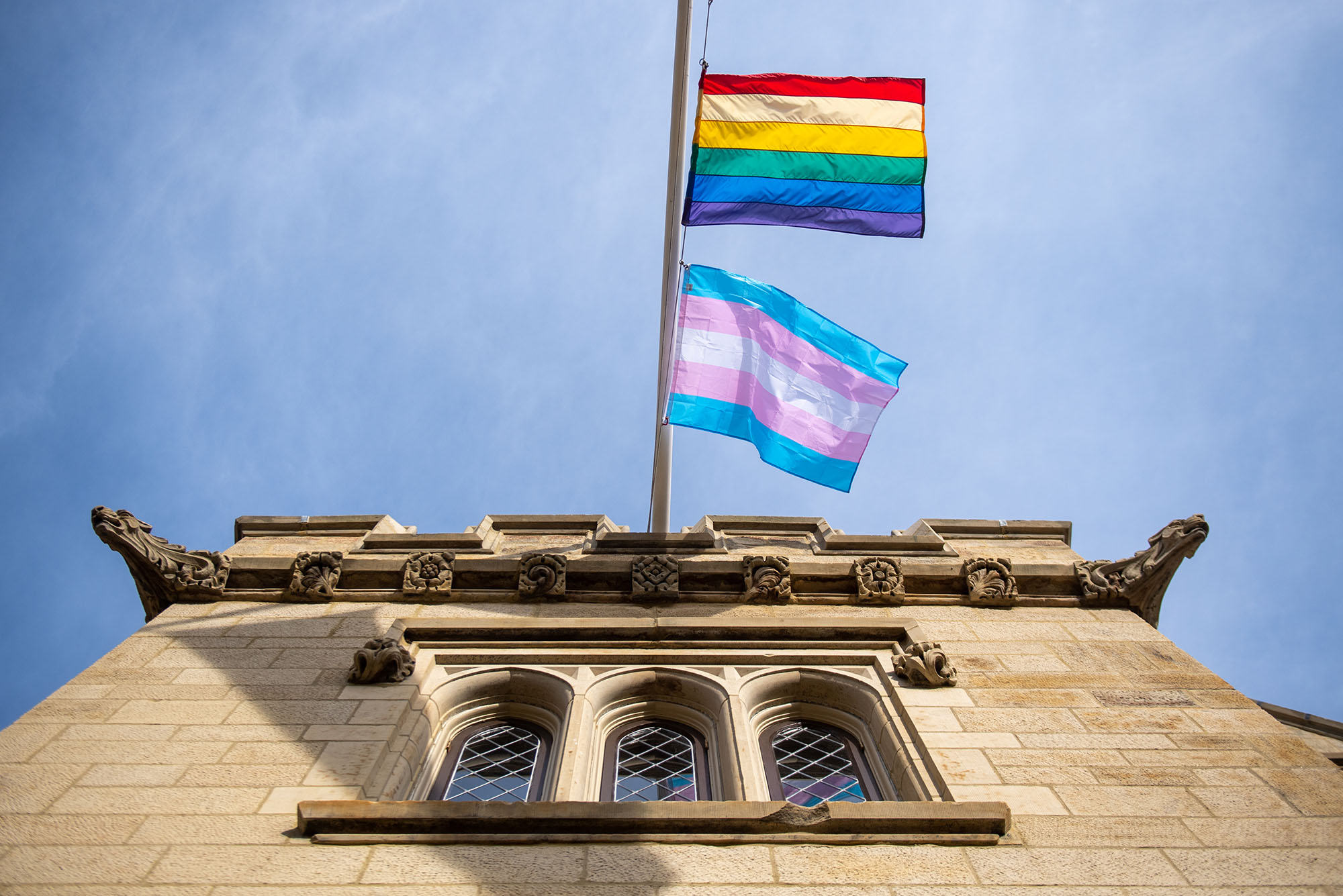 Photo of the Transgender Day of Remembrance flag flying over the Dahod Family Alumni Center. The pride flag (a rainbow flag) and the transgender flag (aa flag of light blue, pink, and white) wave in the wind on a pole attached to a large, tan brick building.