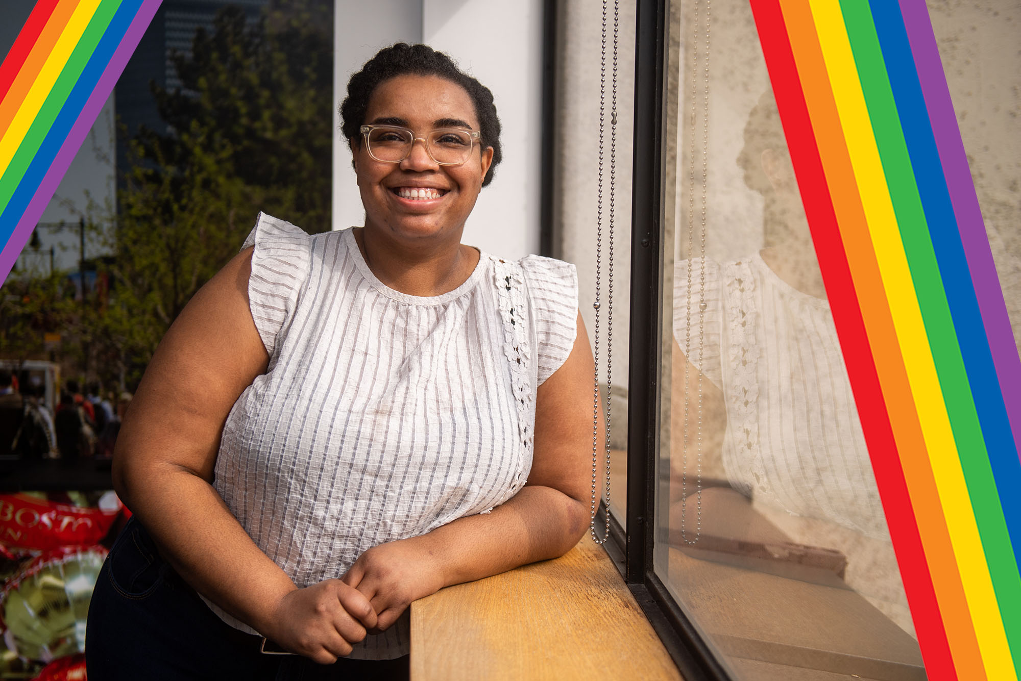 Photo of Chantel James, a young Black woman with clear glasses. She smiles, and wears a white blouse with tufted sleeves. She leans against a window in a room, presumably an office. Small pastel-colored rainbows are overlaid at left and right of the photo.
