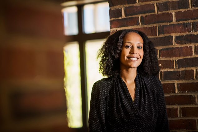 Photo of Clinical Assistant Professor in Health Science, Hagere Yilma, posing for a photo. A Black woman with shoulder-length black hair styled in a twist-out hairstyle wearing a black, tunic-style blouse stands in front of a brick wall and smiles to the camera.