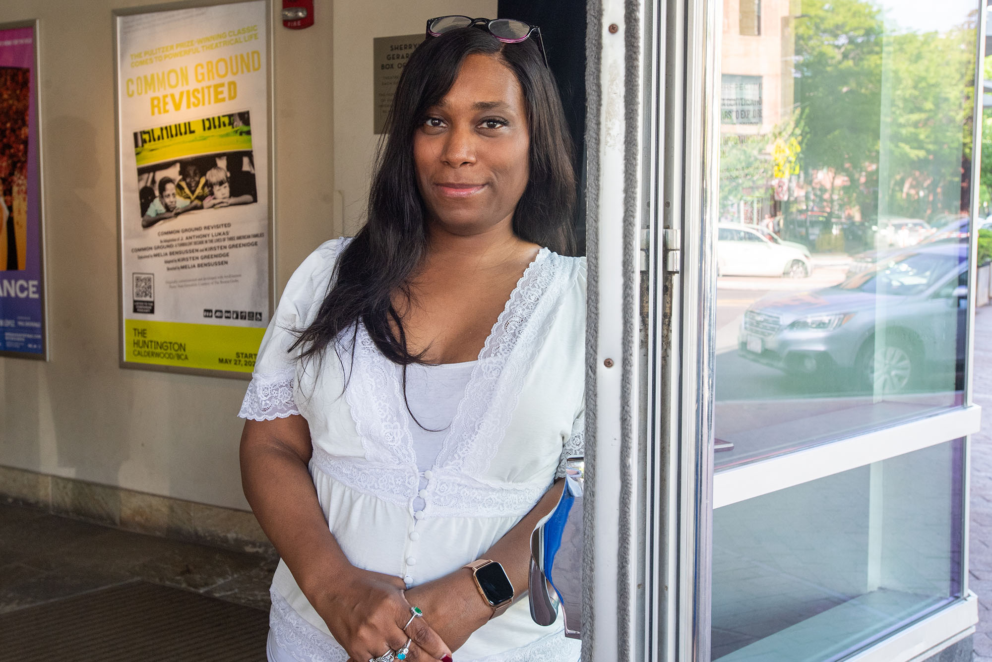 Photo of BU playwright Kirsten Greenidge, aBlack woman with long, straight Black hair. She leans against a door wearing a white blouse as glasses res atop her head. The poster for "Common Ground Revisited" can be seen on the wall behind her.