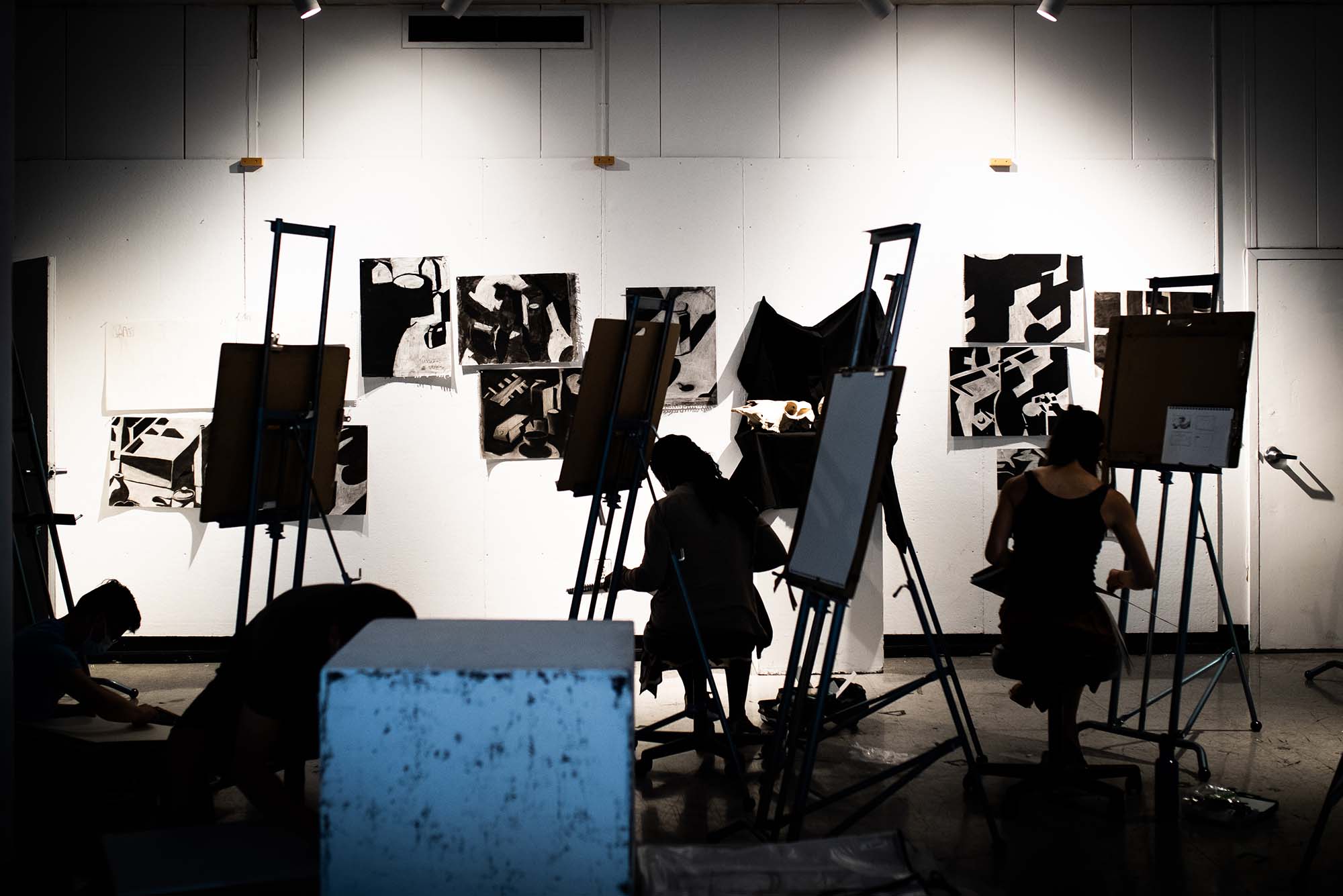 Photo taken from behind students seated at easels as they participate in an Introduction to Drawing class at CFA on June 7, 2022. The are back lit and are seen in silhouette. On a white wall in front of them, large black and white geometric drawings are seen. A white-ish blue box is seen in the left foreground.