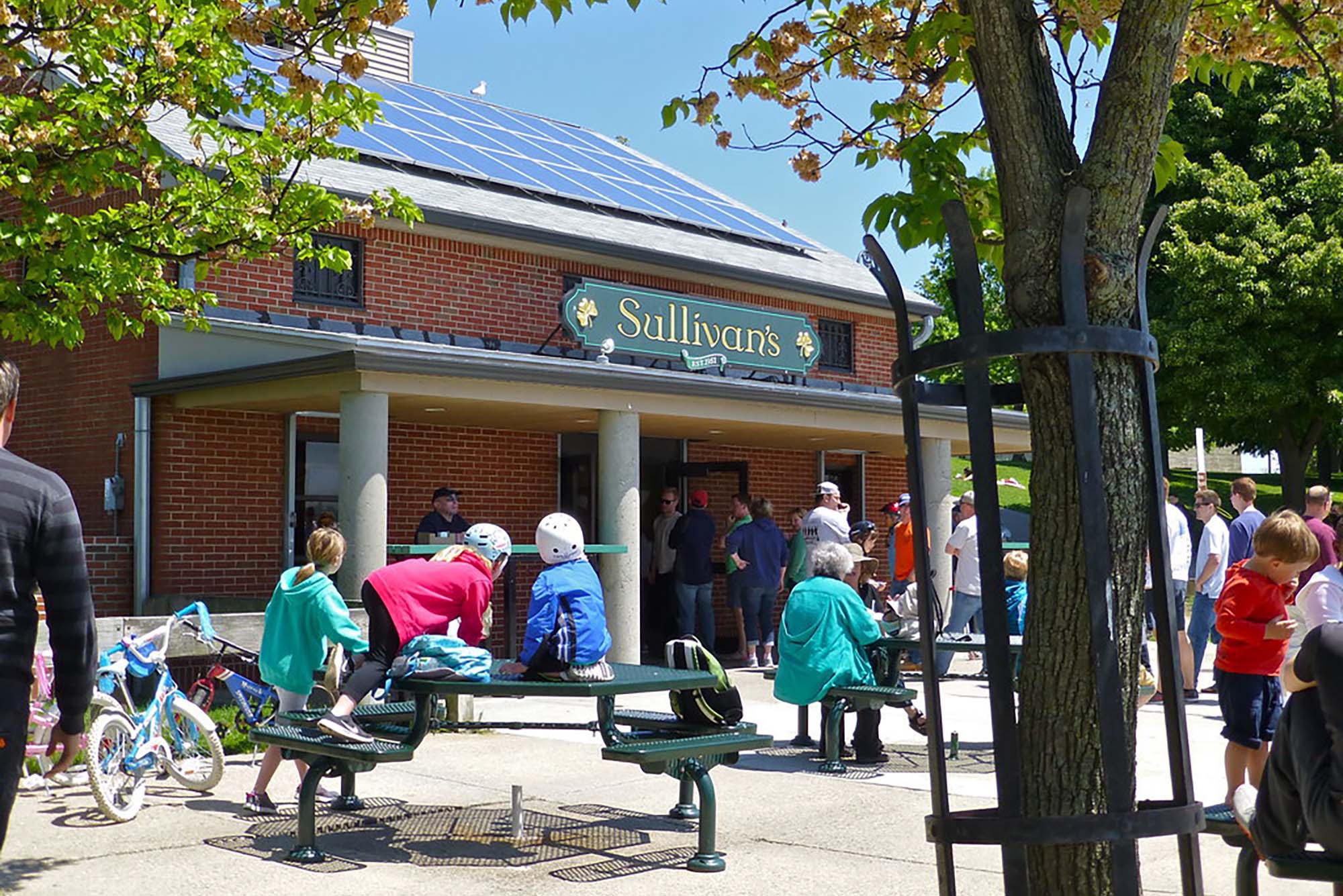 Photo of people gathered in font of a building with a large green sign that reads "Sullivan's". The sit and stand on and around dark green tables on a sunny day.