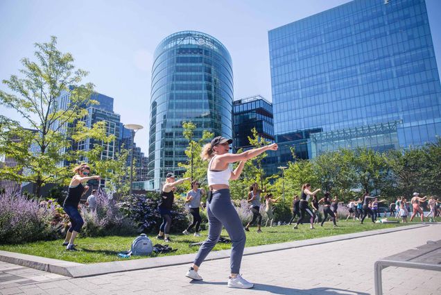 Photo of a women wearing an athletic crop top and leggings punching the air as she speaks into a wireless headset on a bright, sunny day. A group of people in athletic wear can be seen copying her punch as they stand on the green lawn behind her. City buildings can be seen looming in the distance.