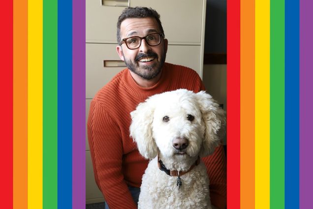 Headshot of Luis Menéndez-Antuña smiling in a red sweater and dark-framed glasses. He poses in front of an office filing cabinet with a white doodle dog. Rainbow red, orange, yellow, green, blue, and purple lines frame the left and right sides of the photo.