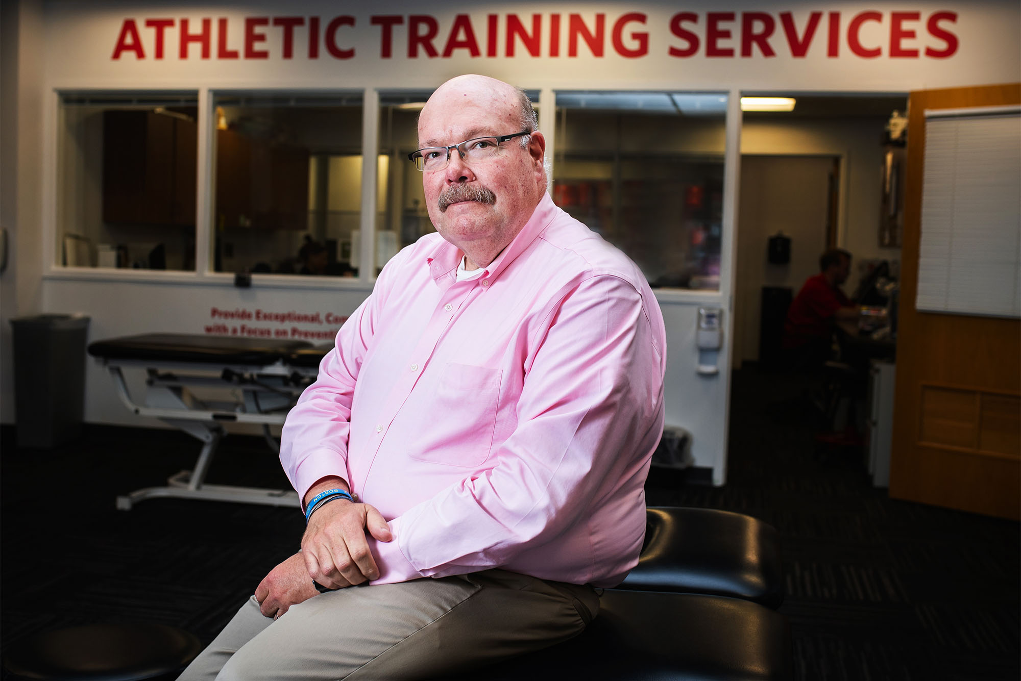 Richard Mark Laursen sits in a white collared shirt underneath a banner reading "Athletic Training Services"