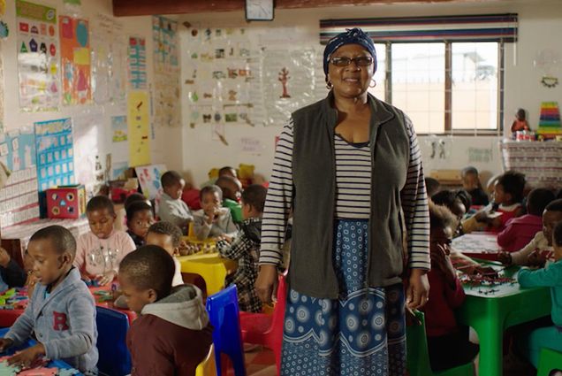 A woman in a striped shirt and vest stands in a classroom full of children, facing the camera