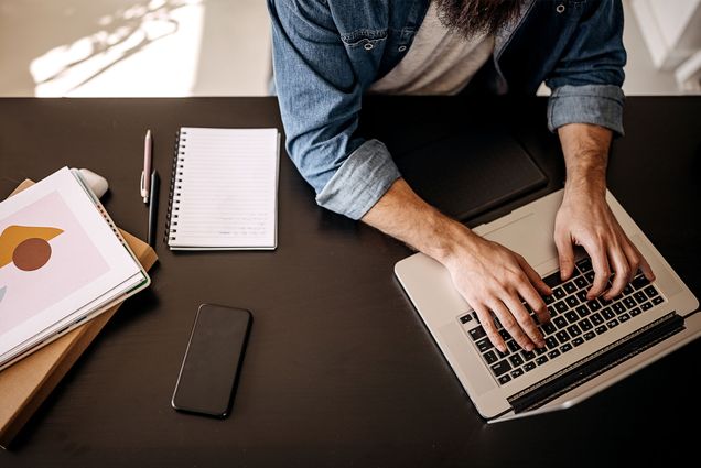 A white man sits at a desk while typing on a laptop. There is a phone and two notebooks nearby. The view is from the top down.