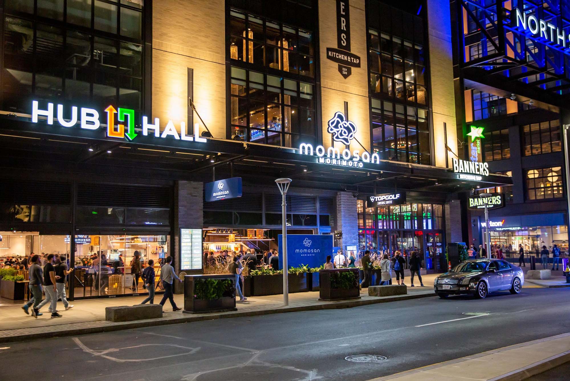 Photo of the facade of Hub Hall taken from Causeway Street in Boston. In the photo, it is night time, and white Hub Hall sign with green and orange arrows are seen on the facade at left. A glowing sign for a restaurant called "Momosan morimoto" is seen at center. At right are signs for Top Golf and Banners. The North Station sign is seen cut off at top right. In the photo, people walk by on the sidewalk and are seen inside the restaurants.
