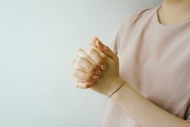 Photo of a person with fingernails painted a glittery pink holds hands in prayer in front of them. Only hands and front of pink shirt are visible, as they stand in front of a pale blue background.