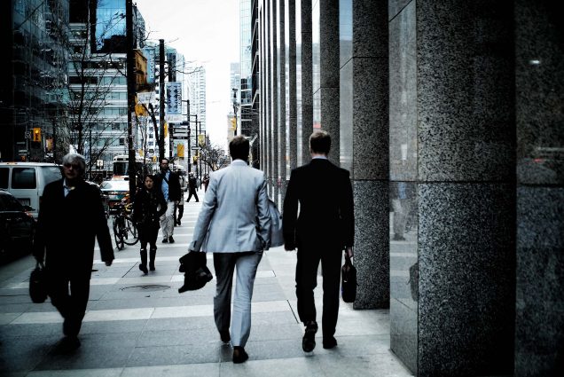Photo of two white men wearing grey and black business suits walking down a busy city street. They both hold black briefcases.