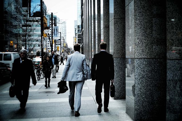 Photo of two white men wearing grey and black business suits walking down a busy city street. They both hold black briefcases.