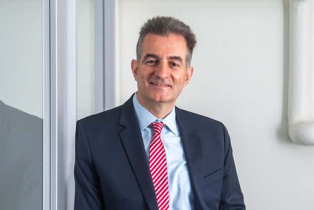 Photo of ENG professor Ioannis Paschalidis, leaning against a glass door and slightly smiling. He wears a dark blue suit, light blue shirt, and red and white striped tie. He is a light-skinned middle-aged man with short brown and gray hair.