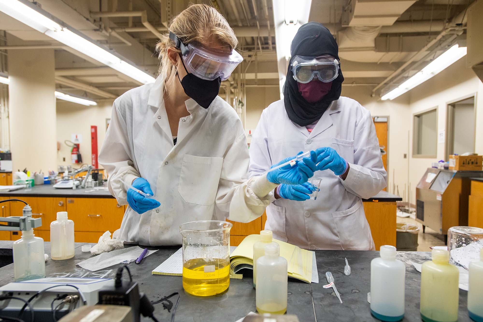 Photo of Gillian Minarik (CAS’23), left, and Nadifa Abdi (CAS’23), working through a pKa of indicators lab during CHEM201, Quantitative Analytical Chemistry Laboratory June 28 in Metcalf Science Hall. Minarik is a White woman with blonde hair hold appears to be passing Abdi a pipette. Abdi is a young woman of color wearing a black hijab. Both wear white lab coats, clear safety goggles, face masks, and blue gloves. On the table in front of them, beakers of yellow liquid are seen.