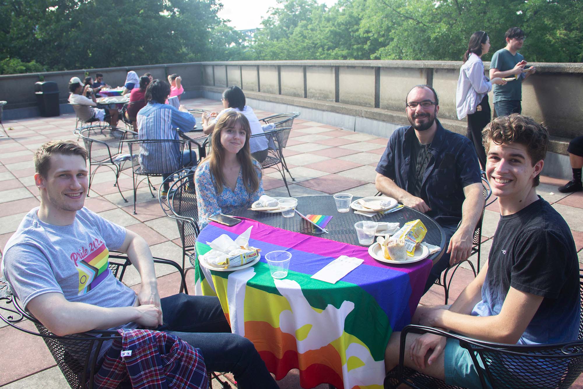 photo of David King (BUMC-IT staff), from left, Lourdes Amieva (MET’22), Scott Amico (BUMC-IT staff), and Shaw Hutton (CGS’25) on the Pride Month BBQ event held by BU Dean of Students’ Office at GSU on Friday, June 24, 2022. They sit at a black metal outdoor table, which has a large, rainbow pride flag draped over the front half. At left, King is a young White man who wears a gray Pride flag t-shirt and smiles. Amieva has long brown hair, light skin and wears a floral blouse as she smiles. Amico wears a dark short sleeve button down and glasses and smiles. Hutton wears a gradient blue t-shirt and has short, curly brown hair and smiles wide. Other folks eating outside at the GSU can be seen behind them.