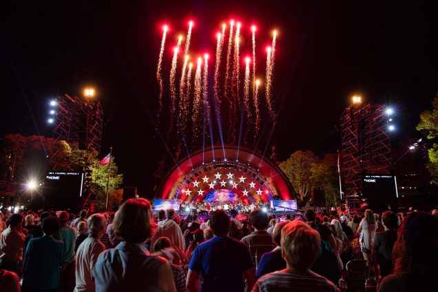 Photo of The Boston Pops performing their annual Fourth of July Fireworks Spectacular at the Hatch Memorial Shell on the Charles River Esplanade. In the photo, the Hatch Shell is lit in red white and blue. The backs of spectators' heads are seen as the look up towards about a dozen fireworks that shoot up from the shell into the black sky.