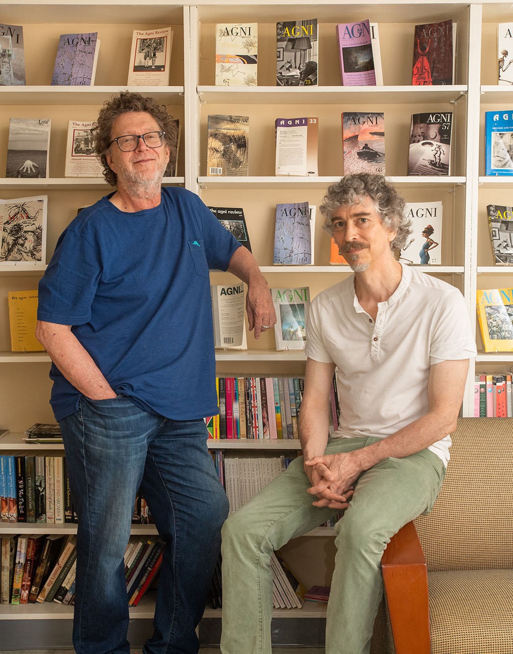 Photo of Sven Birkerts, left, and William Pierce in the AGNI offices on Bay State Rd. Birkerts, a white man with light brown curly hair and a silver grey beard, wears a blue shirt and jeans as he leans back with one arm on a bookcase behind him. Pierce, a white man with grey and brown curly hair wearing a white short sleeved shirt and light green pants, sits on an armchair to the right of Birkerts, also in front of the bookcase. The bookcase behind spans the entire wall and is made of a light wood. Many books on it feature the title "AGNI"