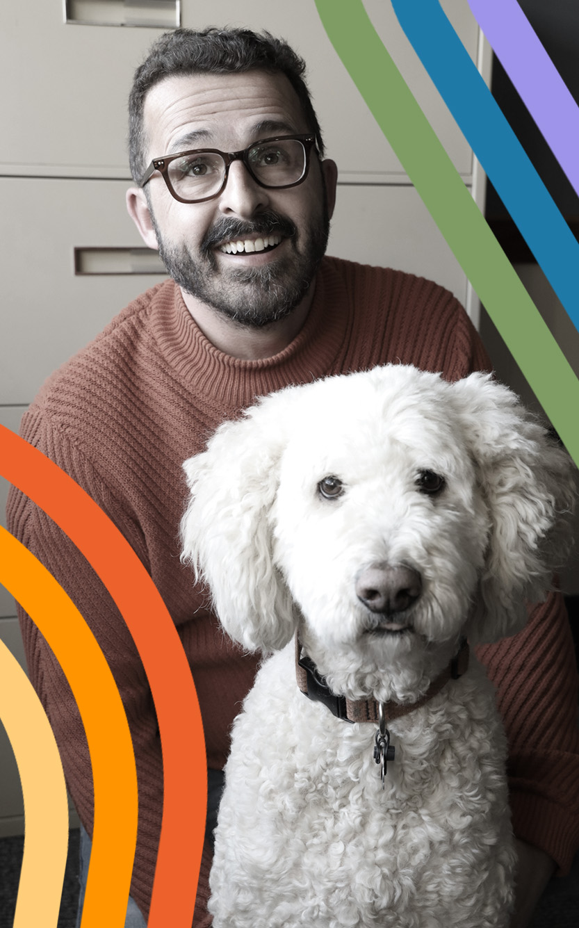 Faded black and white headshot of Luis Menéndez-Antuña smiling in a light red sweater and dark-framed glasses. He poses in front of an office filing cabinet with a white doodle dog. Rainbow red, orange, yellow, green, blue and purple lines can be seen framing the lower left and upper right corners of the photo.