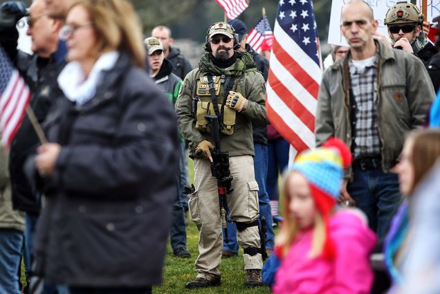 Photo of, at center, a White man with sunglasses, a beard, and wearing a green hat with a military style vest and rifle. He stands amidst a crowd of people and American flags and also has a military style headset. A man at right who is partially seen seems to be wearing similar combat style gear. Children and families can be seen in the ground.