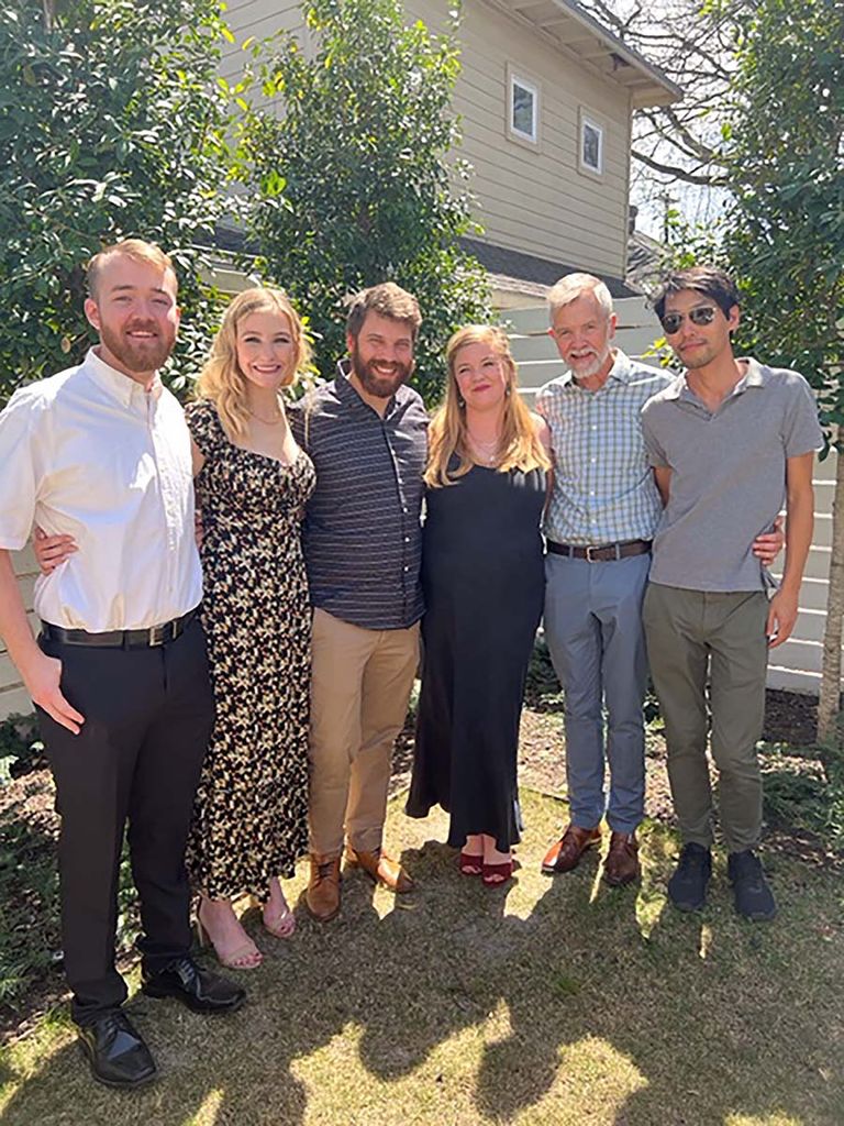 Photo of Chard and his family: son James (from left), daughter Madeleine, son-in-law Samuel, daughter Cecilia, Chard, and husband, Michael, wearing sunglasses. They are all light-skinned and pose in a half circle with their arms around each other. They wear semi-formal clothing and seem to be standing in a backyard. 