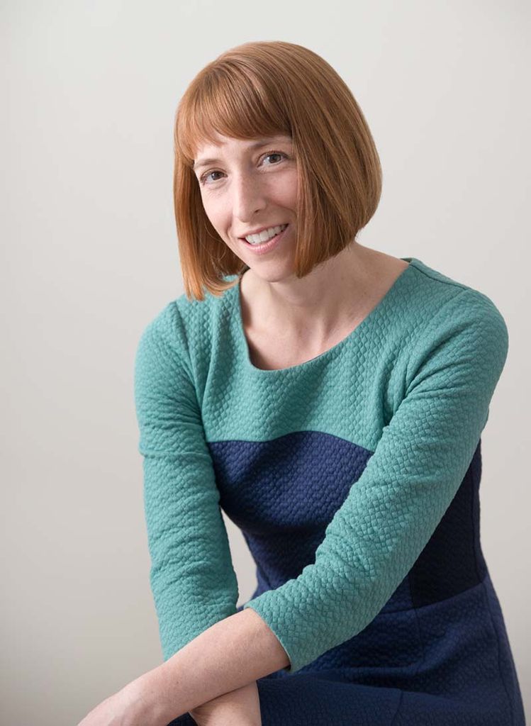 Portrait of Japonica Brown-Saracino, a White woman with short ginger hair who half smiles as she sits and rests her arms on her knee. She wears a teal and blue knit dress. 