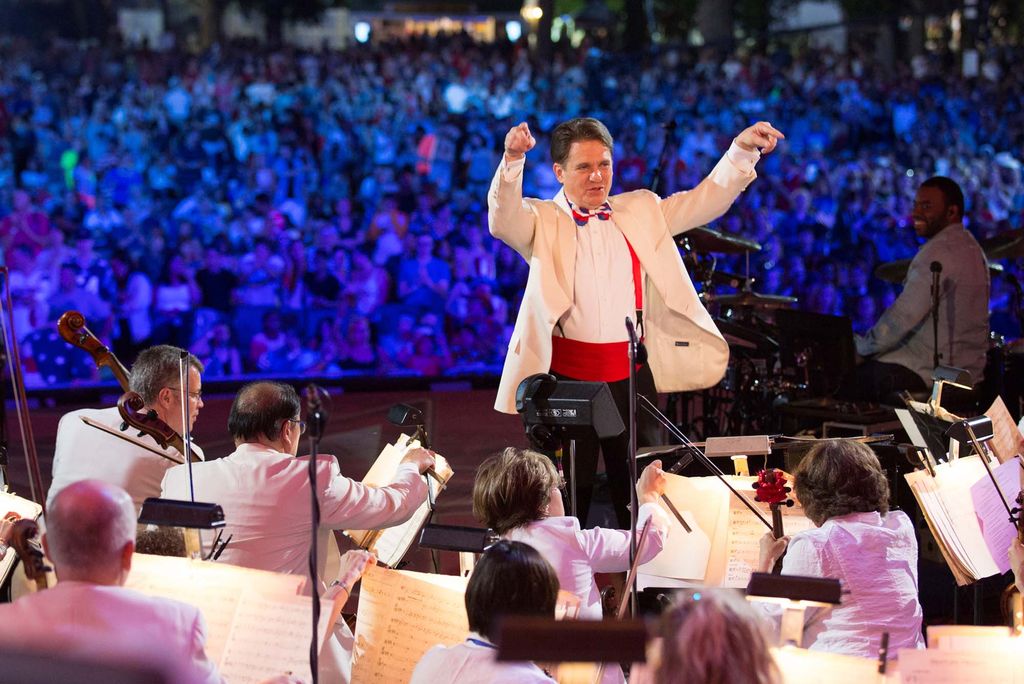 Photo of Maestro Keith Lockhart (Hon.’04) conducting the Boston Pops. He wears a white suit jacket and bright red cummerbund and gestures with his hands raised as he conducts the performers. The back of the performers' heads are seen in the foreground; they sit in front of music stands. Behind them, a crowd, lit in blue, is seen along the Esplanade.