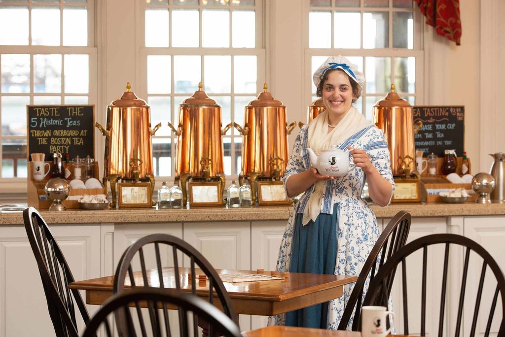 Photo of a White woman dressed in a white bonnet and blue, floral dress standing with a white teapot in front of a series of large, gold tea carafes. A wooden table and chairs is seen in the foreground.