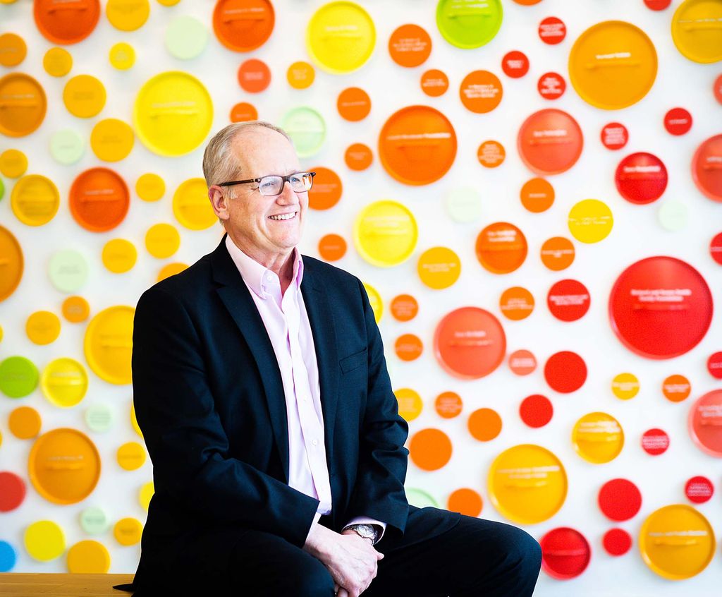 Photo of David Water, an older white man wearing black glasses, black blazer and pants, and a pastel pink collared shirt. He sits on a stool and looks tot he right of the camera. He smiles in front of a background featuring bright red, yellow, and orange dots on a white background.