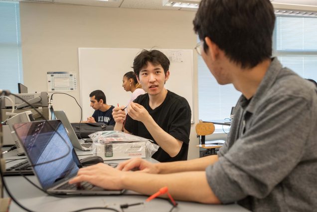 Photo of Young Chan Cho (ENG’24), left, and Jung Won “Ed” Park (ENG’24) during an electrical engineering lab called Thevenin Equivalence Oscilloscopes in Photonics June 15. Cho is a young AAPI man and wears a black t-shirt and holds two wires in his hand as he looks across the table towards Park. Park wears a gray button down with rolled up sleeves and clear glasses and types at his laptop as he looks towards Cho. other students working on the lab can be seen behind him.
