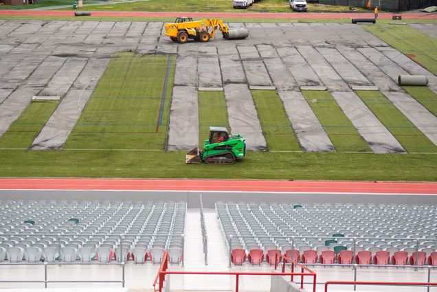 Photo of a bright green bobcat machine and a larger, yellow machine with a large roller hanging off an arm, traversing Nickerson field. Large rolls of the astroturf being pulled off can be seen running North-South up the field. In the foreground, the gray bleachers of Nickerson are seen.