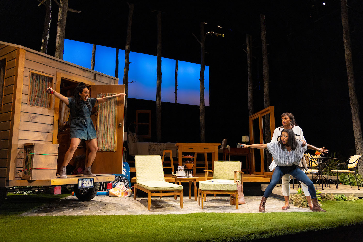 Photo of Lyndsay Allyn Cox (left), Arie Thompson and Nikkole Salter in The Huntington Theatre production of "Our Daughters, Like Pillars" by Kirsten Greenidge which ran earlier this season.The stage is set up to look like a camping scene, with two beach chairs in the middle and an open "house" to the left. Cox stands in the house with arms raised in glee, while Thompson and Salter stand and crouch to the right of the stage as they point at Cox in astonishment.