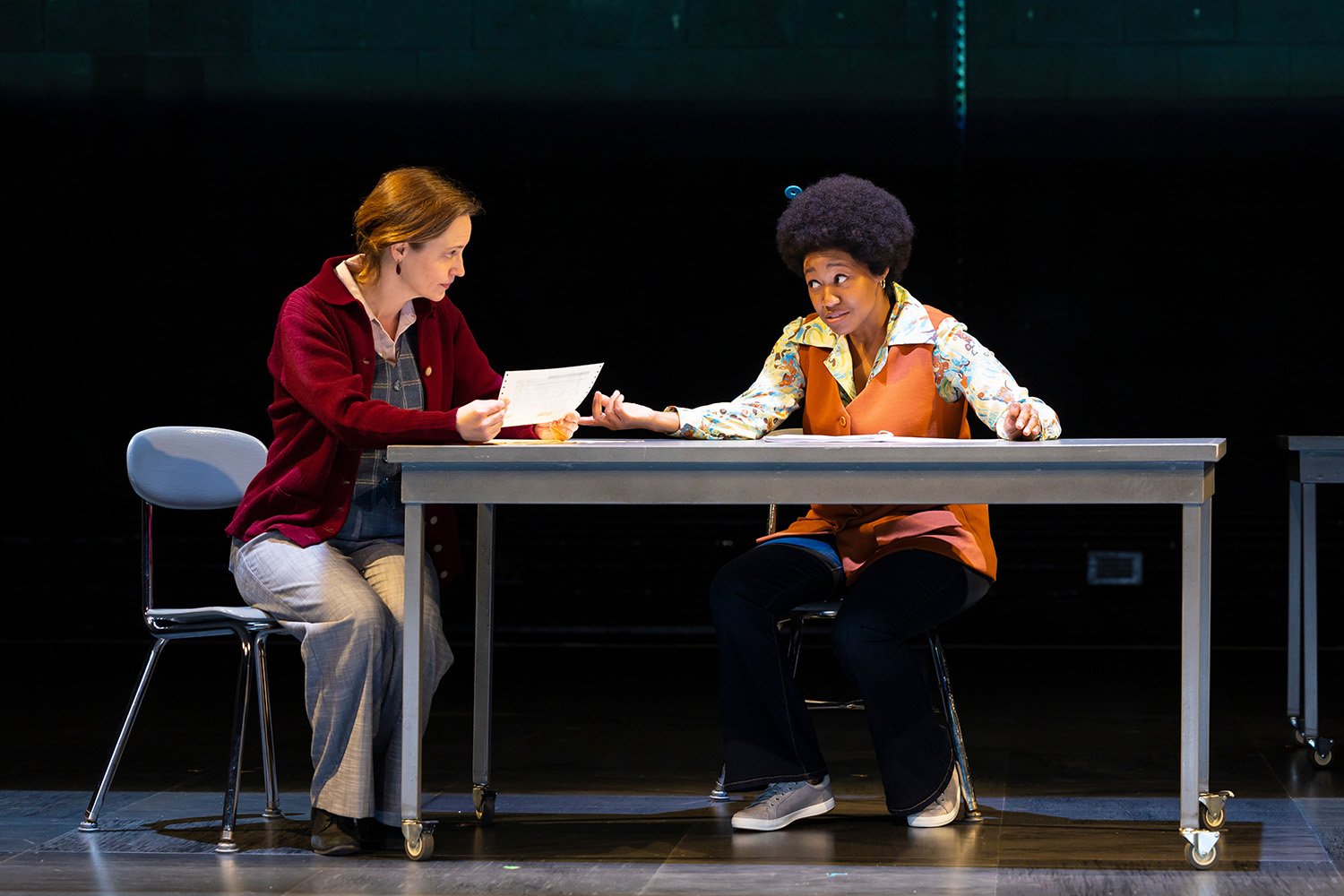 Photo of Stacy Fischer (left) and Elle Borders in a scene from "Common Ground Revisited" at The Huntington. Fischer has her red hair tied back and wears a red cardigan, plaid shirt, and grey slack as she sits in a chair on the left side of a table in the middle of the stage. She holds a piece of paper in her hands. Borders sits to the right of Fischer, in the middle of the table. Her her is styled in an afro with a small blue pick in it, and she wears a floral-patterned collared shirt, burnt orange dress overtop the shirt, and jeans.