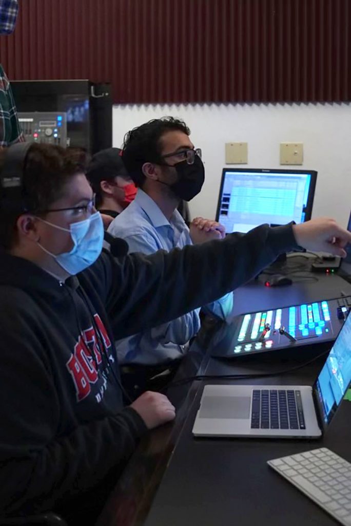 Photo of Jacob Cohen (COM‘22) (left) and Evan Jimenez (COM’23) in the BUTV10 control room. Both young men wear masks and glasses. Cohen wears a blue BU sweatshirt and points to something out of frame with his left hand with his laptop open in front of him. Jimenez wears a blue button-down shirt and sits in front of a control board. 
